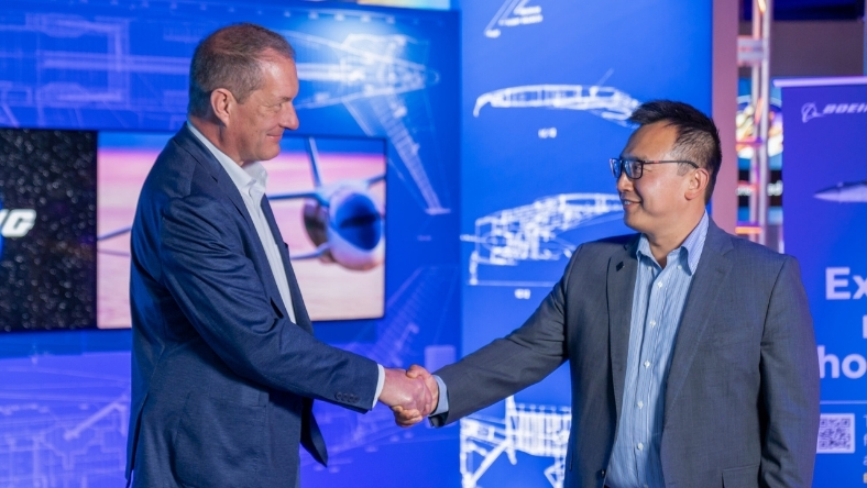 Two aerospace executives shake hands before display boards showing aircraft design diagrams